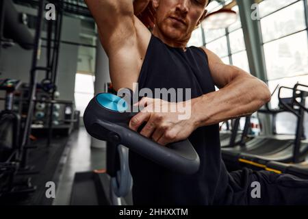 Ein junger Mann benutzt und genießt die Schlagmassagepistole, um sich zu entspannen und die Schmerzen im Körper und in den Muskeln im Fitnessstudio zu lindern. Stockfoto