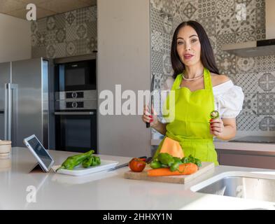 Trendige Brünette in Schürze hält Messer und ein Stück Pfeffer Blick auf die Kamera, während die Zubereitung gesunder Salat in der Küche zu Hause Stockfoto