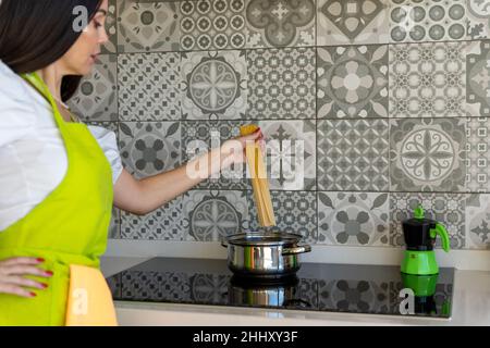 Trendige Brünette in Schürze, die Hand an der Taille hält und Spaghetti in den Topf legt, während sie in der Küche zu Hause das Mittagessen kocht Stockfoto