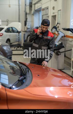 Ein junger, selbstbewusster Mechaniker, der mit dem Kunden auf dem Mobiltelefon spricht, während er nach der Überprüfung und Reparatur mit dem Auto steht Stockfoto