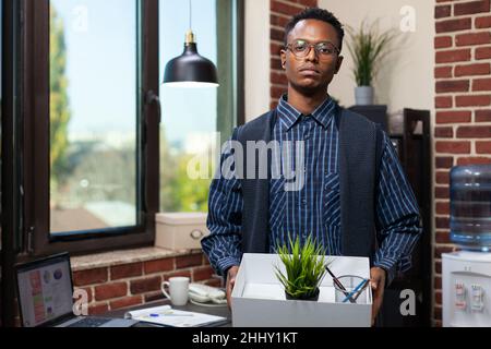 Porträt enttäuscht aussehender afroamerikanischer Mitarbeiter, der nach der Entlassung das Büro mit persönlichen Gegenständen verlassen hat. Entlassen Startup-Arbeiter verlieren Job nach Unternehmen verkleinerten. Stockfoto