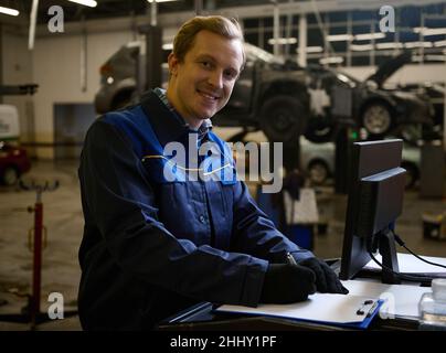 Konzentrierter Auto-Mechaniker Techniker Autoingenieur lächelt Blick auf Kamera schreiben Checkliste auf Zwischenablage und arbeiten auf Monitor während der Wartung einer Ca Stockfoto