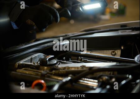 Nahaufnahme der Hände eines Automechanikers, der den Motor eines Autos mit einer leuchtenden Taschenlampe inspiziert. Stockfoto