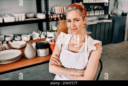 Ich lebe mein bestes Leben. Beschnittenes Porträt einer attraktiven reifen Frau, die mit gefalteten Armen in ihrer Töpferwerkstatt sitzt. Stockfoto