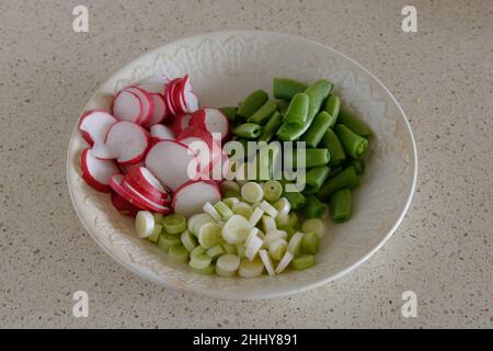 Frische Salatzutaten - Rettich, Frühlingszwiebeln, Zuckererbsen, die in einer weißen Schüssel auf beigem Hintergrund gehackt wurden Stockfoto