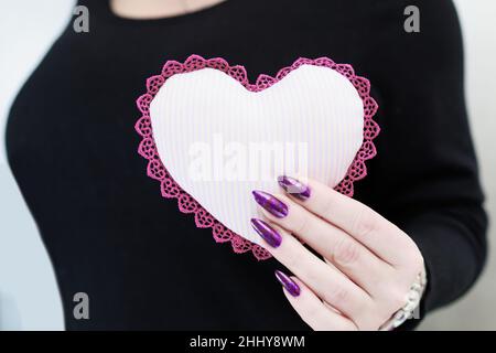 Weibliche Hände mit lila langen Nägeln halten ein weiß rosa Herz Stockfoto