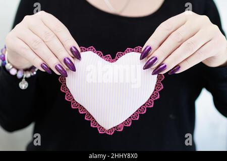 Weibliche Hände mit lila langen Nägeln halten ein weiß rosa Herz Stockfoto