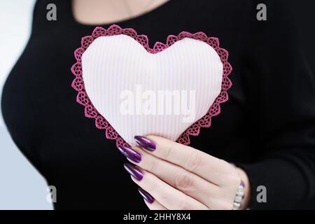 Weibliche Hände mit lila langen Nägeln halten ein weiß rosa Herz Stockfoto
