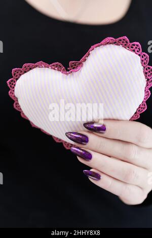Weibliche Hände mit lila langen Nägeln halten ein weiß rosa Herz Stockfoto