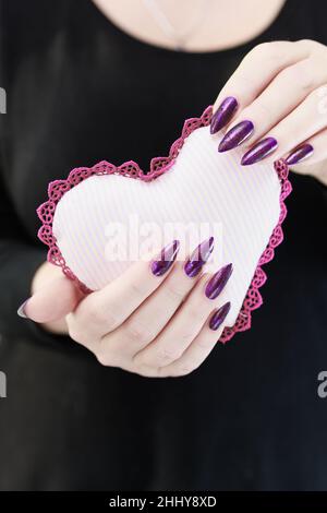 Weibliche Hände mit lila langen Nägeln halten ein weiß rosa Herz Stockfoto