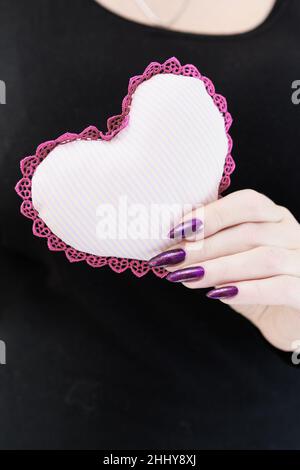 Weibliche Hände mit lila langen Nägeln halten ein weiß rosa Herz Stockfoto