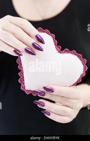 Weibliche Hände mit lila langen Nägeln halten ein weiß rosa Herz Stockfoto