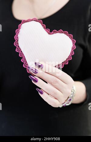 Weibliche Hände mit lila langen Nägeln halten ein weiß rosa Herz Stockfoto