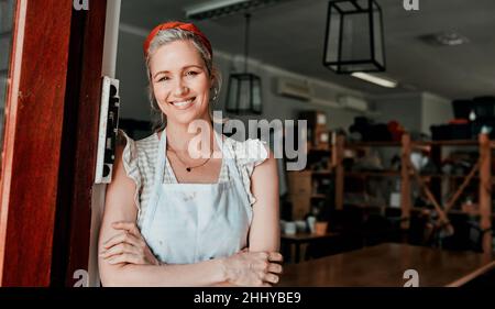 Bitte, Kommen Sie herein. Beschnittenes Porträt einer attraktiven reifen Frau, die mit gefalteten Armen in ihrer Töpferwerkstatt steht. Stockfoto