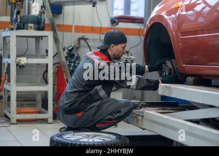 Moderne Werkstatt oder Techniker in der Arbeitskleidung Wechselrad des Autos, während sitzen auf Kniebeugen vor dem Auto Stockfoto