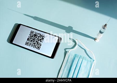 Smartphone mit Quar-Code des Impfzertifikats auf dem Bildschirm, Spritze und medizinischer Maske auf hellblauem Hintergrund. Stockfoto