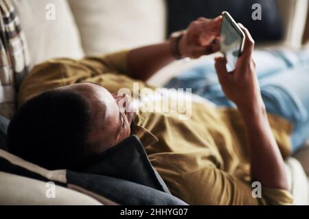 Zeit für Social Media. Eine kurze Aufnahme eines hübschen jungen Mannes, der ein Smartphone benutzt, während er sich zu Hause auf seiner Couch entspannt. Stockfoto