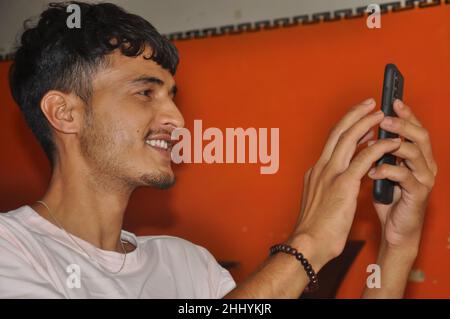 Nahaufnahme eines jungen Mannes, der lächelt, während er sein Telefon benutzt, während er drinnen sitzt Stockfoto