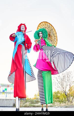 Die ganze Länge der kreativen talentierten Darsteller mit Gesichtskunst in bunten Kostümen und Perücken, die an sonnigen Tagen auf Stelzen im Park mit asiatischem Regenschirm und Schwert während der Show stehen Stockfoto