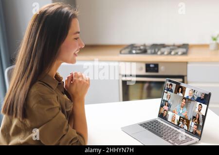 Remote-Meeting per Videoanruf. Asiatische Frau, die in der Küche sitzt und die App auf dem Laptop für die Online-Kommunikation mit einer Gruppe verschiedener multirassischer Menschen nutzt. Rückansicht Stockfoto