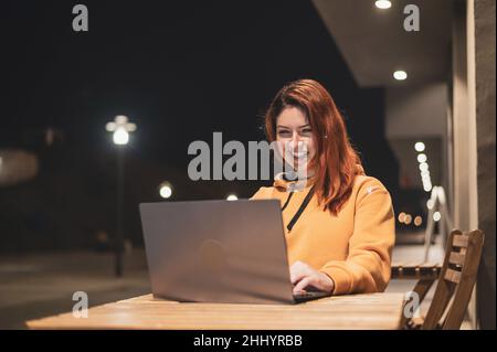 Eine Frau arbeitet am späten Abend in einem Sommercafé an einem Laptop. Happy Girl studiert, während sie auf einer leeren Straße an einem Holztisch sitzt Stockfoto
