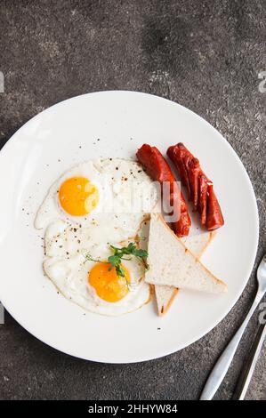 Spiegeleier und Würstchen werden auf einem weißen Teller auf grauem Hintergrund serviert. Stockfoto