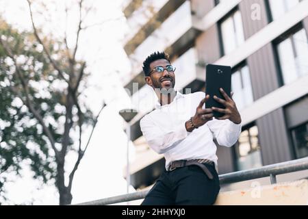 Lächelnder junger schwarzer Geschäftsmann in weißem Hemd, der ein Foto mit seinem Tablet auf der Straße machte Stockfoto