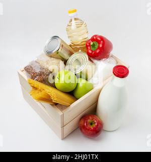 Schachtel mit Lebensmittelspenden. Äpfel, Spaghetti, Zucker, Krups, Sonnenblumenöl, Bulgarischer Pfeffer, Konserven, Milch in einer Holzkiste auf weißem Hintergrund Stockfoto