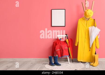 Stuhl und Gestell mit Regenmänteln, Regenschirmen, Gummistiefeln an der rosafarbenen Wand Stockfoto