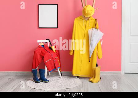 Stuhl und Gestell mit Regenmänteln, Regenschirmen, Gummistiefeln an der rosafarbenen Wand Stockfoto