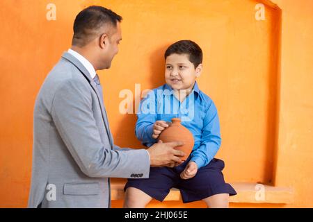 indischer Mann mit kleinem Kind, der Münze in eine Tongeldkiste legt.männlicher Bankmanager und kleiner Junge, der traditionelle Sparschweine oder Gulak hält und lehrt, zu sparen Stockfoto