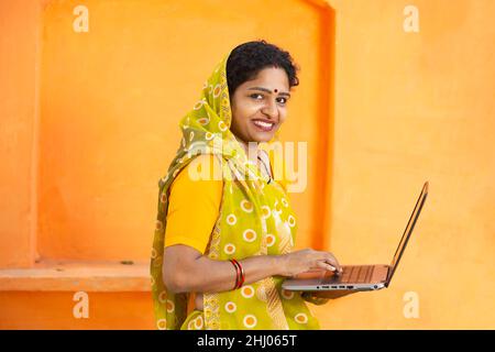 Porträt einer glücklichen jungen indischen Frau mit Laptop. Lächelnde Frau trägt Sari und hält den Computer vor orangefarbenem Hintergrund. Sie schaut in die Kamera, s Stockfoto