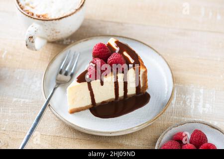 Käsekuchen mit Schokoladensauce und Himbeeren auf einem Teller, Holztisch Hintergrund. Serviert mit einer Tasse Cappuccino-Kaffee Stockfoto