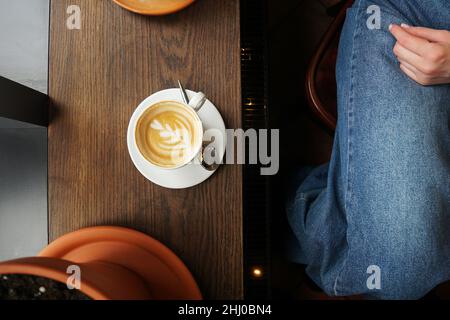 Einfache Portion Cappuccino-Kaffee mit herzförmiger Blumenkunst auf dunklem Holztisch. Blaue Jeans eines Mädchens, das in einem Café sitzt Stockfoto