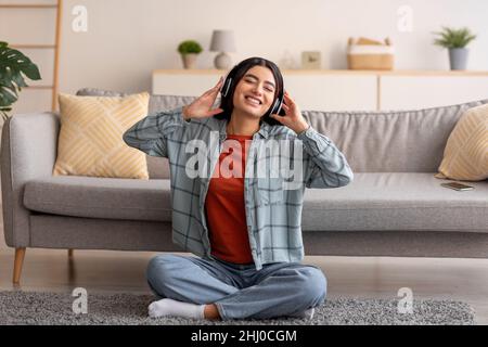 Schöne junge arabische Frau, die auf dem Boden sitzt, Kopfhörer trägt und zu Hause Musik hört Stockfoto