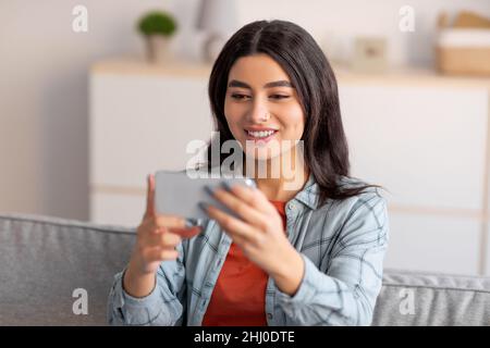 Schöne junge arabische Frau, die mit dem Smartphone auf der Couch sitzt, Filme gucken, von zu Hause aus arbeitet oder studiert Stockfoto