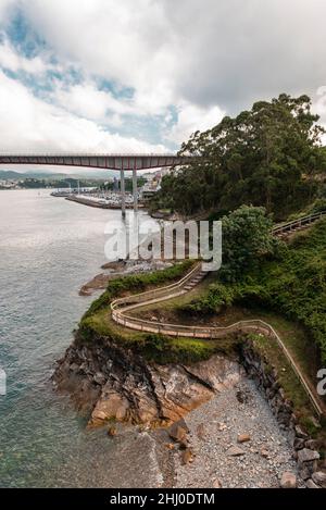 Im Vordergrund mit baumgesäumter Küste und Holzfußbrücke und im Hintergrund die Stadt, der Hafen und die Brücke über die Mündung. Stockfoto