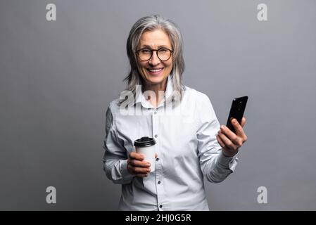 Charmante ältere Frau in formeller Kleidung stehen mit Papierbecher heißes Getränk und Smartphone isoliert auf grau, Vorbereitung auf einen guten Arbeitstag. Reife Geschäftsfrau macht eine Kaffeepause Stockfoto