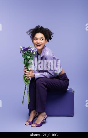 Die ganze Länge der glücklichen afroamerikanischen Frau sitzt auf dem Koffer und hält Blumen auf lila Stockfoto