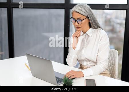 Entspannte ältere Frauen sitzen bequem am Tisch im Büro, schreiben E-Mail auf Laptop, schreiben Nachricht, arbeiten an Projekt. Stock Foto Stockfoto