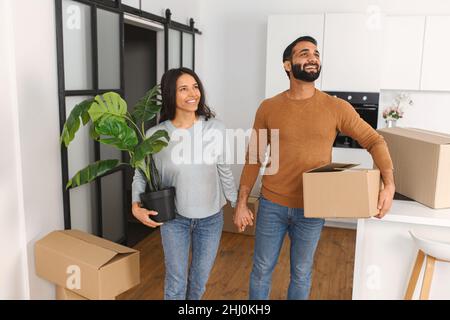 Glückliches Paar, das sich nach dem Umzug mit dem neuen Zuhause wunderte. Glücklicher Mann, der Pappkarton trägt und sich an eine Frau festhält, die das Werk hält. Immobilienmiete, Hypothek und Eigentumskonzept Stockfoto