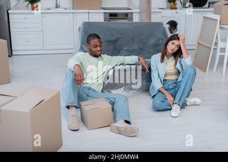Unzufriedenes multiethnisches Paar, das in Plastikfolie neben Boxen und Sofa sitzt Stockfoto