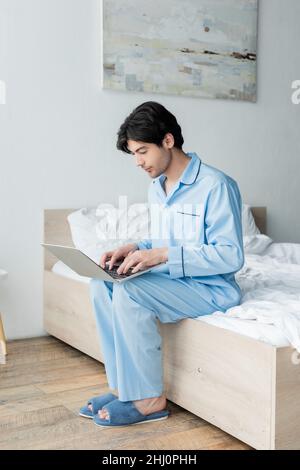 Mann in blauem Schlafanzug und Pantoffeln sitzt auf dem Bett und tippt auf dem Laptop Stockfoto