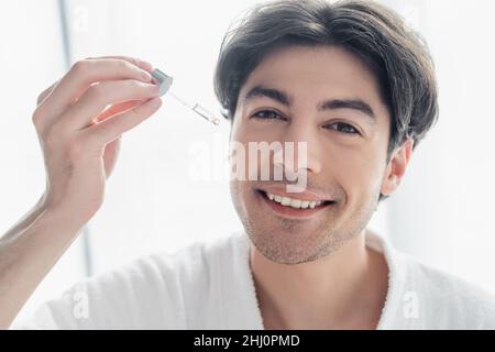 Glücklicher Mann, der die Kamera ansah, während er kosmetisches Serum im Badezimmer applyte Stockfoto