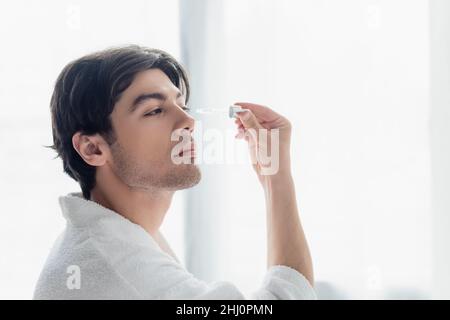 Junger Mann im weißen Bademantel, der kosmetisches Serum im Badezimmer anwendet Stockfoto