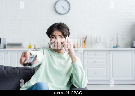 Fröhlicher Mann, der auf dem Smartphone spricht, während er zu Hause fernsieht Stockfoto