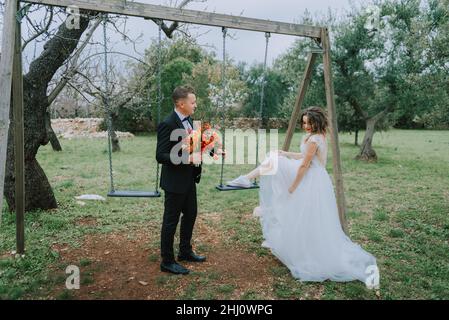 Fröhliches, stilvolles und lächelndes Paar, das am Hochzeitstag in der Toskana, Italien, unterwegs ist. FRISCH VERHEIRATET MIT DER SCHAUKEL IM PARK. Die Braut und der Bräutigam gehen den hinunter Stockfoto
