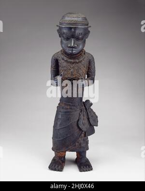 Männliche Figur: Hofbeamter der Edo-Völker des 16th.–17th. Jahrhunderts Gelehrte haben vorgeschlagen, dass diese Figur auf einem Gedenkaltar platziert wurde, der einem König oder oba von Benin gewidmet ist. Die Figur ist mit einem markanten Kreuzanhänger dargestellt. Sein Wickelrock ist mit Profilköpfen portugiesischer Händler, einem frontalen afrikanischen Kopf und anderen gängigen Benin-Motiven wie Flussblättern, Mudfish- und Interlace-Mustern geschmückt.für diese Figur wurden drei verschiedene Identitäten vorgeschlagen. Der Beamte kann ein Bote eines Herrschers sein, der als „Ogane“ bezeichnet wird, der heute als Anführer oder „oni“ bezeichnet wird Stockfoto