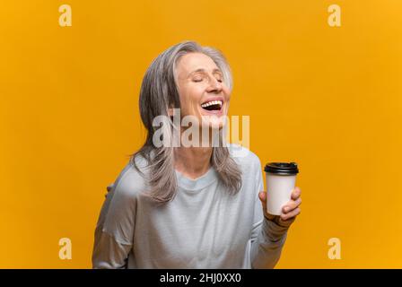 Lächelnde ältere Frau in legerer Kleidung, die eine Papiertasse mit Kaffee isoliert auf gelbem Hintergrund hält. Reife, glückliche, grauhaarige Dame, die steht und lacht, während sie Kaffee zum Ausgehen genießt Stockfoto