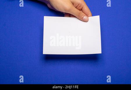 Weibliche Hand hält ein leeres Stück Papier mit Kopierraum vor einem blauen Hintergrundkarton Stockfoto